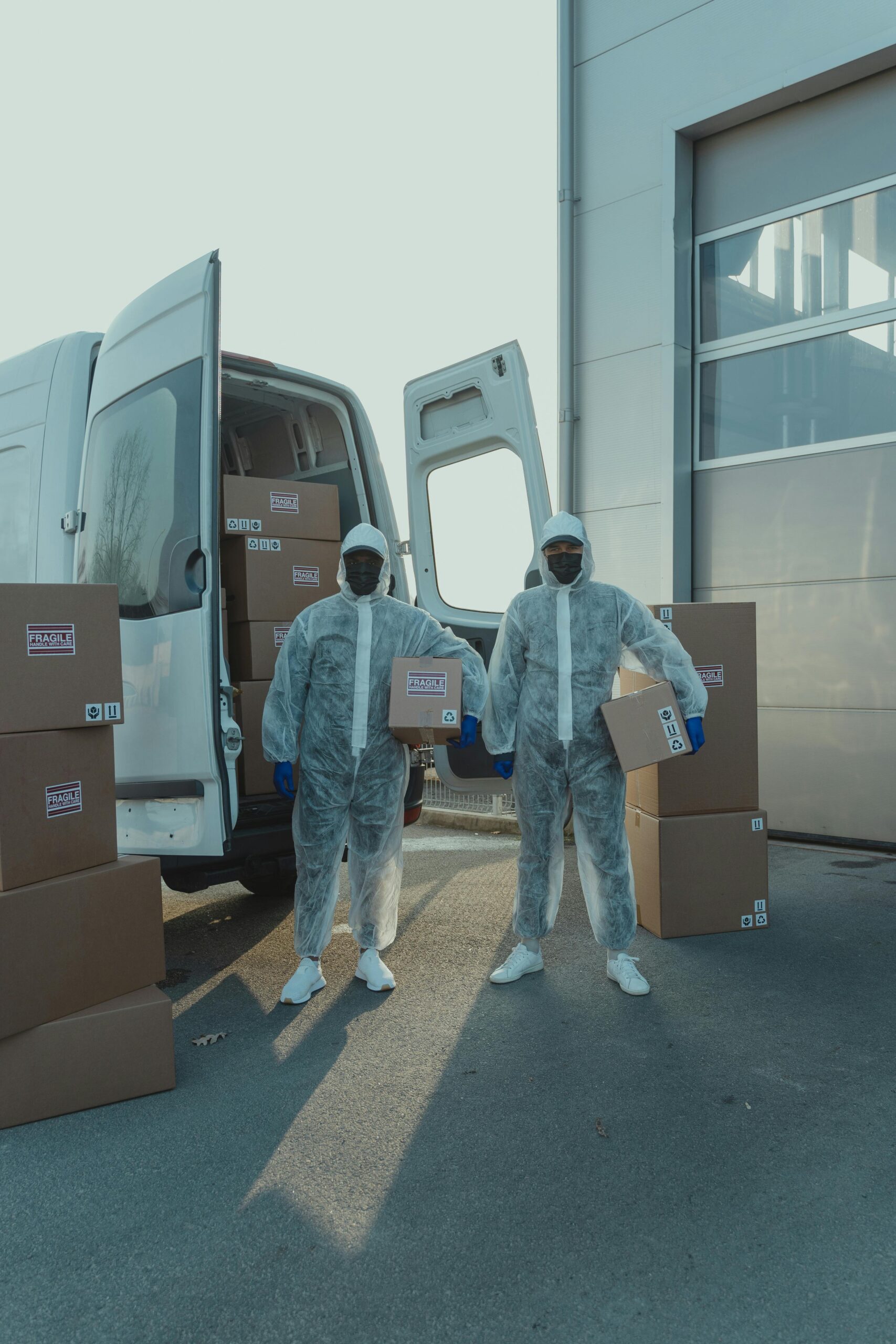 Home Two workers in protective suits load cardboard boxes into a van outdoors.